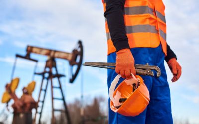 Cropped close-up snapshot of a man wearing blue overalls orange vest and gloves, holding a pipe wrench and a helmet on foreground, oil pump jack on background. Concept of petroleum industry.